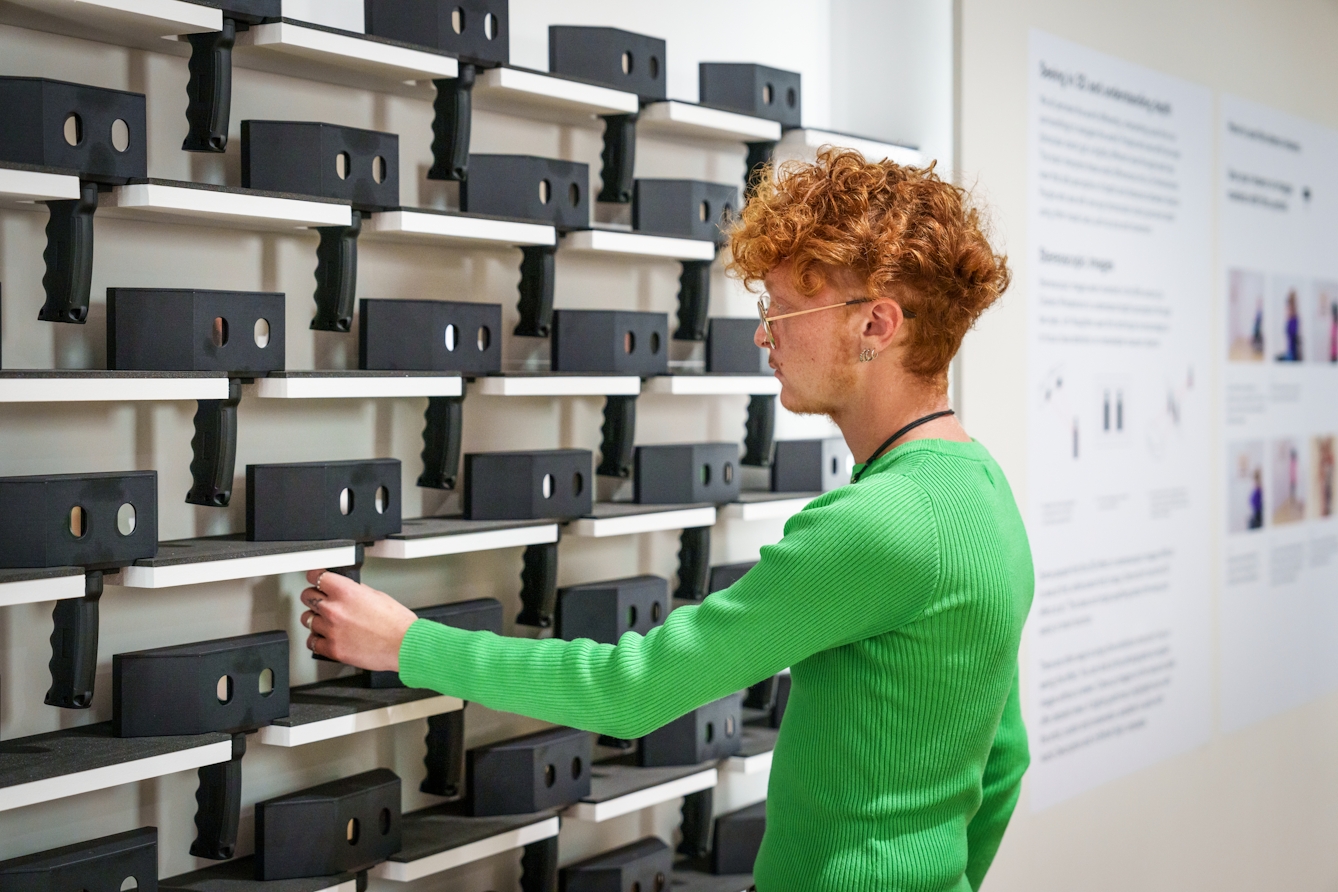 Photograph of a gallery exhibition space showing a visitor wearing a green jumper about to take a stereoscopic viewer from a rack of wall shelving which contains many more viewers.