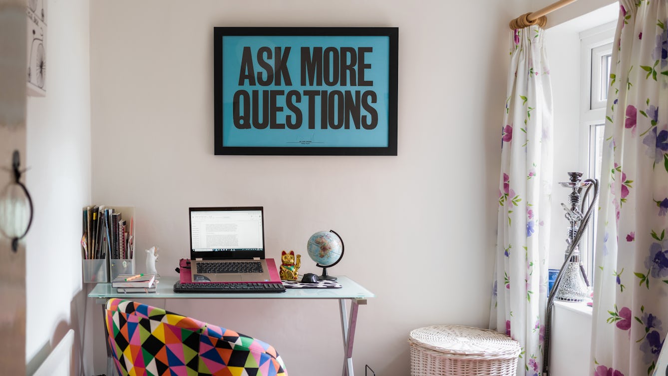 Photograph of a room in a house showing a small desk area with a laptop open on a stand. Next to the laptop is a small globe, note books and file tidies. In front of the desk is a colourful triangular patterned material covered chair. To the right of the desk is a wicker laundry bin, a window with floral curtains and a shisha on the window sill. Hung on the wall above the desk is a large framed picture with a blue background and large black capital letters with the words, 'ASK MORE QUESTIONS'.
