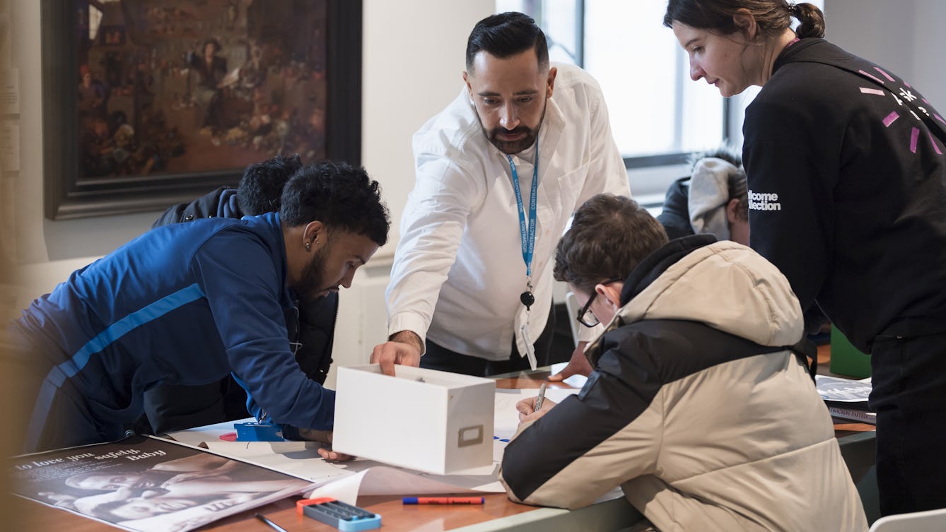 A photograph showing teenage students sitting down and leaning over a table, on which there are pens, posters and other materials. A male adult supervisor with a staff lanyard and shite shirt is place a box down at one end of the table. A female staff member wearing a Wellcome Collection t shirt is leaning over the students' work by the table. 