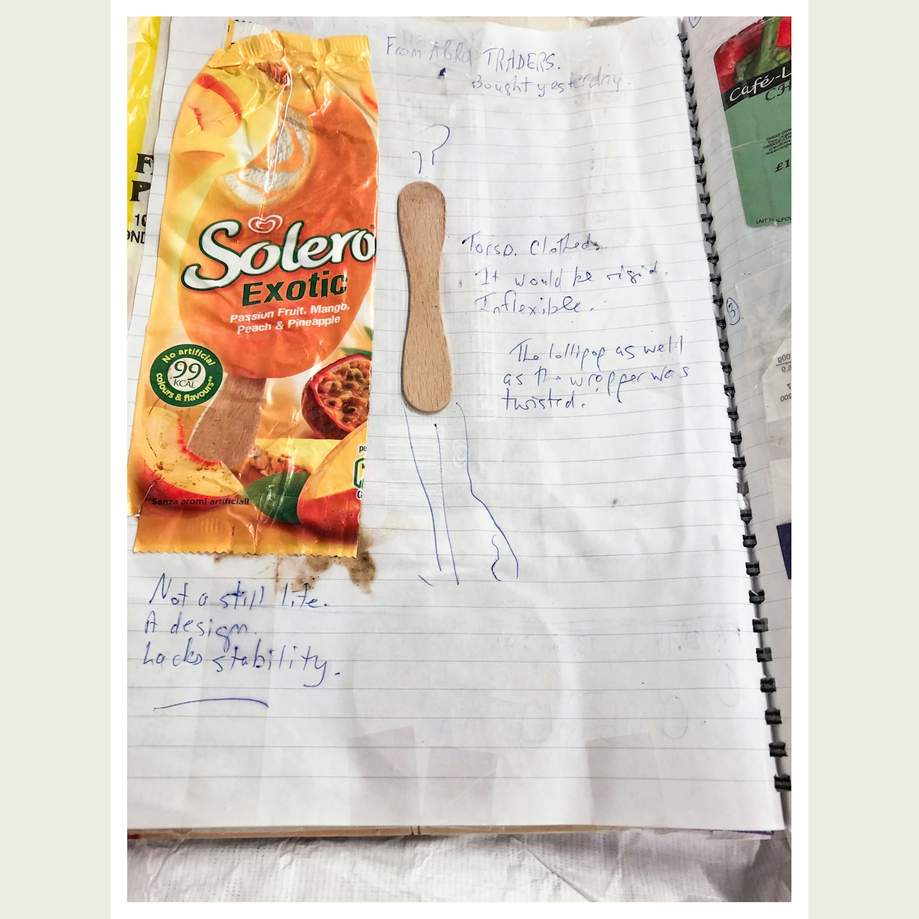 Photograph showing a page from an open lined scrapbook. Stuck to the page is a Solar wrapper and curved wooden ice lolly stick. A drawing around the sticks adds a head and legs to it, making it into a human figure. Handwritten on the page are the words, 'From Abra Traders. Bought yesterday. Torso. Clothed. It would be riding. Inflexible. The Lollipop as well as the wrapper was twisted. Not a still life. A design lacks stability.'