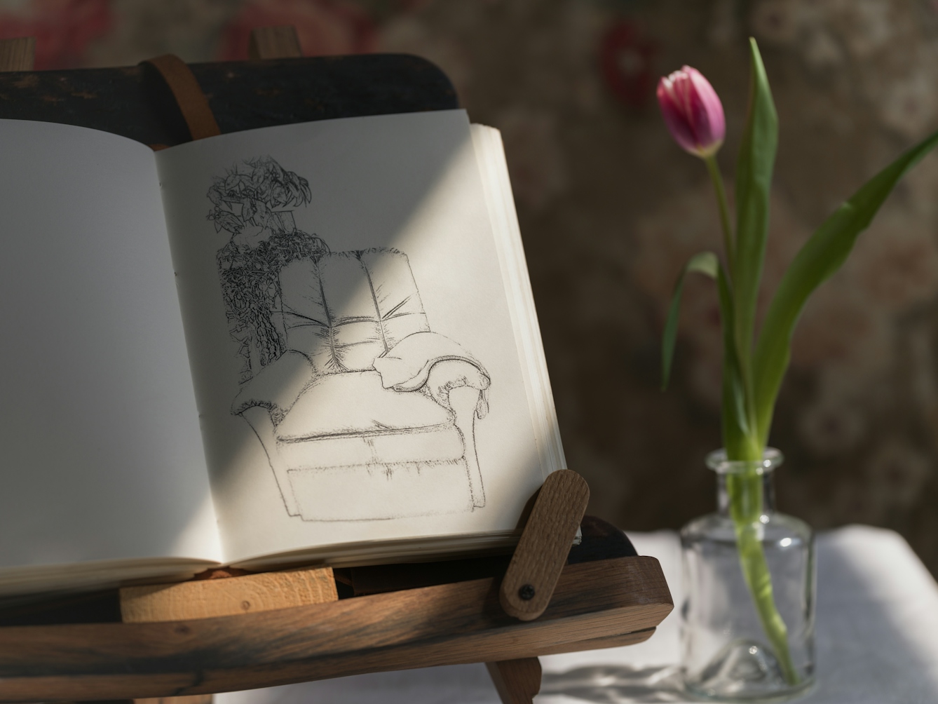 Photograph of a notebook on a wooden book stand, lit by a window from the right, casting a shadow on half of the page. On the right page there is a sketch of an empty armchair. To the right of the book a single tulip in a small glass vase.
