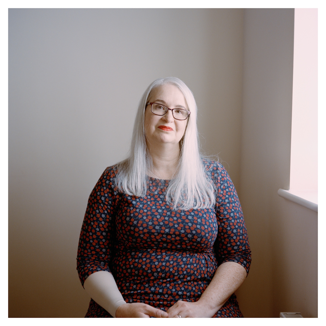 Portrait photograph of Louise, a woman with long grey hair. She is sitting down and looking directly at the camera with a neutral expression.
She is wearing black rectangular glasses, red lipstick and a blue and red floral patterned dress. Her hands are clasped on her lap. There is a white wall behind her, and a window ledge.