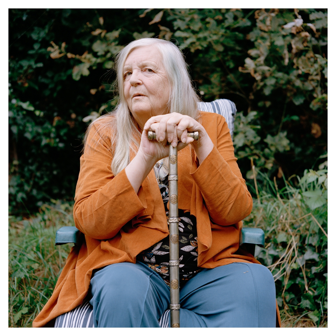 Portrait photograph of Alison, a woman with long grey hair, sitting on a plastic garden chair. Her hands are both resting on a silver walking stick, one hand stacked on top of the other. She is wearing blue trousers, a patterned top and an orange jacket. She has a neutral expression and is looking at the camera. Behind her are are some trees and small plants.