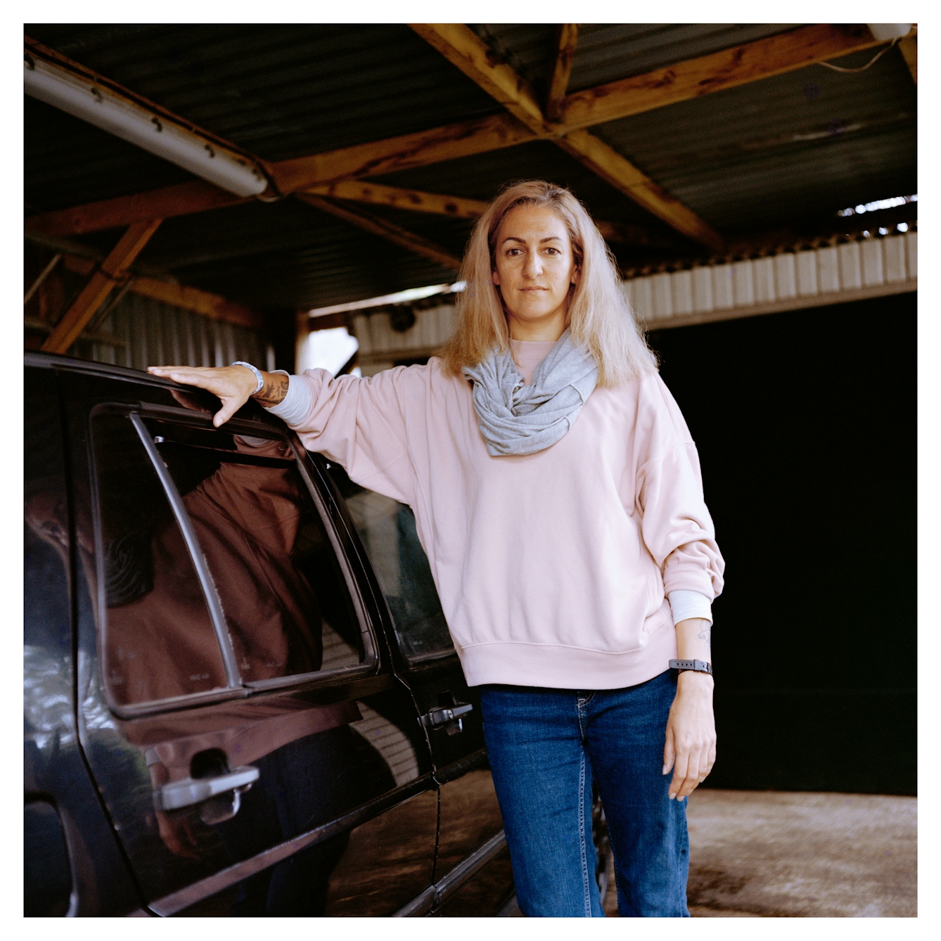 Portrait photograph of Hannah, a woman with shoulder-length blonde hair, leaning against a black car in a garage. She is wearing a pink sweatshirt, a grey scarf and blue jeans. She is looking directly at the camera with a slight smile.
