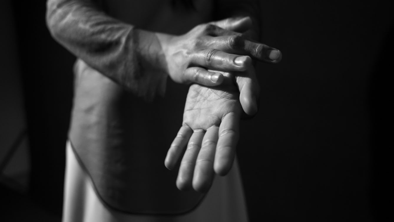 Black and white film still of a performance showing a close-up of an individual gentle rubbing their hands together in a caring manor.