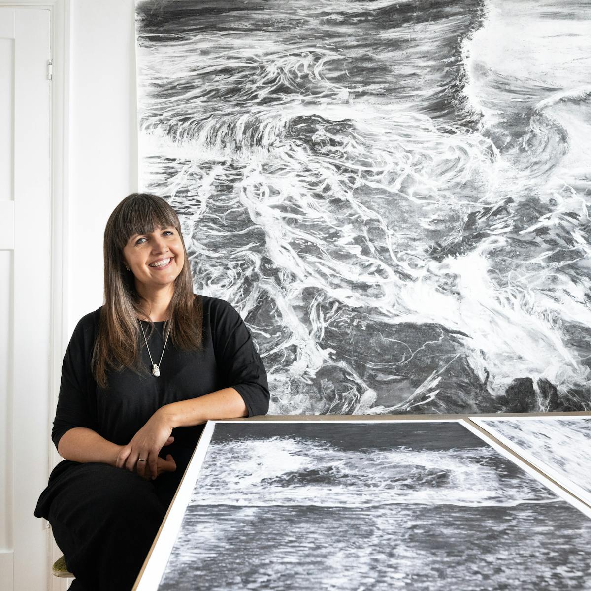 A photograph of the artist Liz Atkin wearing all black seated at a table, surrounded by large scale charcoal works on paper.