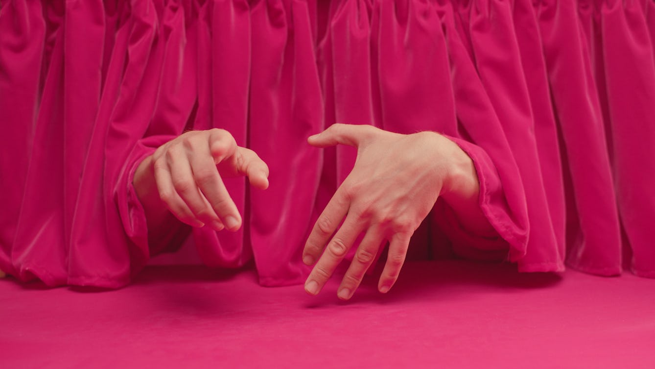 A pair of disembodied hands emerge from a heavy velvet curtain, gesticulating.