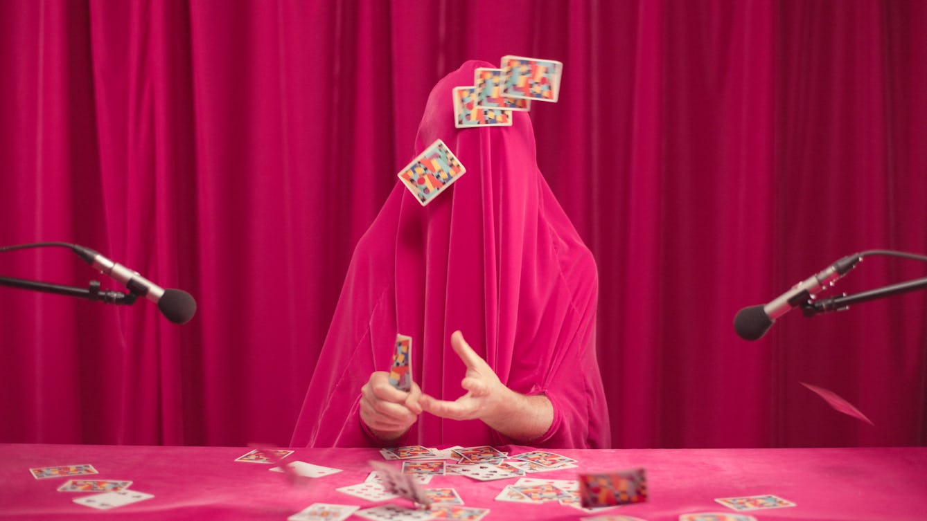 A person shrouded in a pink sheet of material sits at a table, in front of a heavy curtain of the same colour. Only their gesticulating hands are visible. Scattered playing cards lie on the table in front of them. Four cards are also stuck to the sheet covering the head and the figure holds one card in their hand. On either side of the figure is a microphone, directed down towards the hands. 